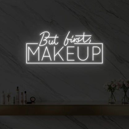 Photo of the 'But first, MAKEUP' neon sign glowing in cool white against a styled interior wall.