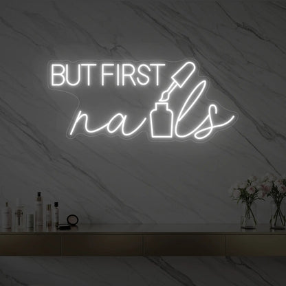 Photo of the 'BUT FIRST nails' neon sign with a nail polish bottle graphic glowing in cool white against a styled interior wall.
