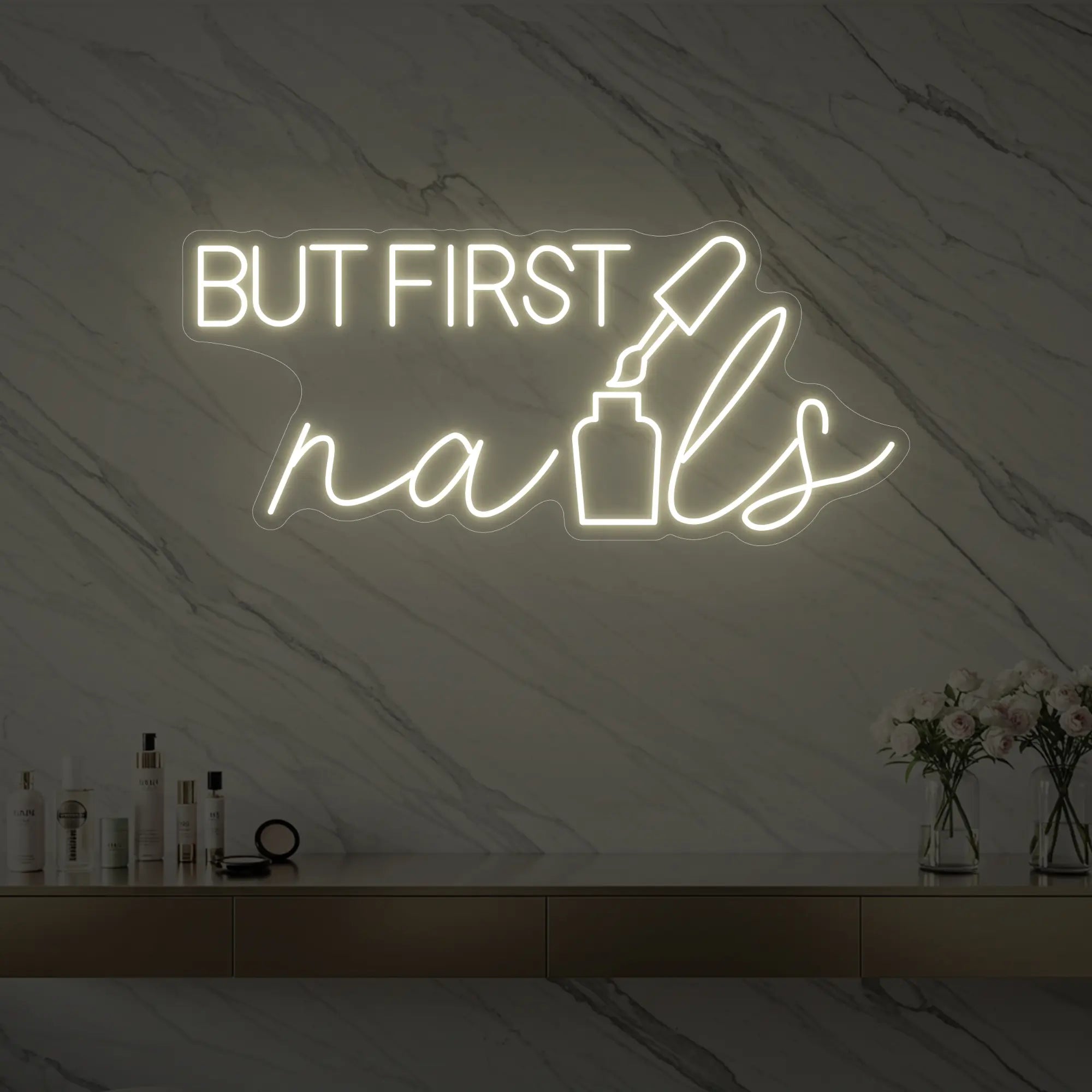 Photo of the 'BUT FIRST nails' neon sign with a nail polish bottle graphic glowing in warm white against a styled interior wall.