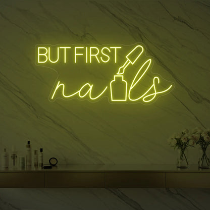 Photo of the 'BUT FIRST nails' neon sign with a nail polish bottle graphic glowing in lemon yellow against a styled interior wall.