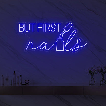 Photo of the 'BUT FIRST nails' neon sign with a nail polish bottle graphic glowing in deep blue against a styled interior wall.
