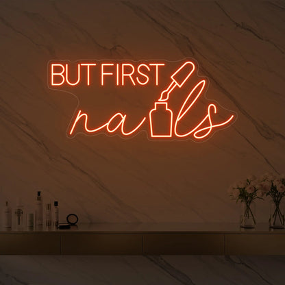 Photo of the 'BUT FIRST nails' neon sign with a nail polish bottle graphic glowing in golden yellow against a styled interior wall.