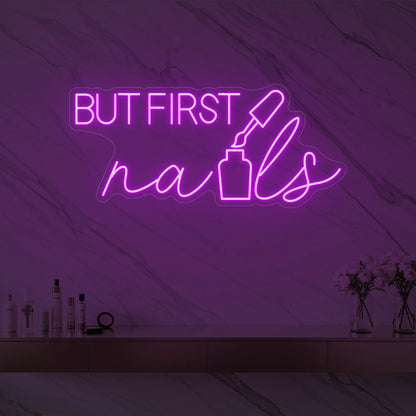 Photo of the 'BUT FIRST nails' neon sign with a nail polish bottle graphic glowing in deep pink against a styled interior wall.