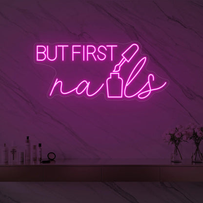 Photo of the 'BUT FIRST nails' neon sign with a nail polish bottle graphic glowing in light pink against a styled interior wall.