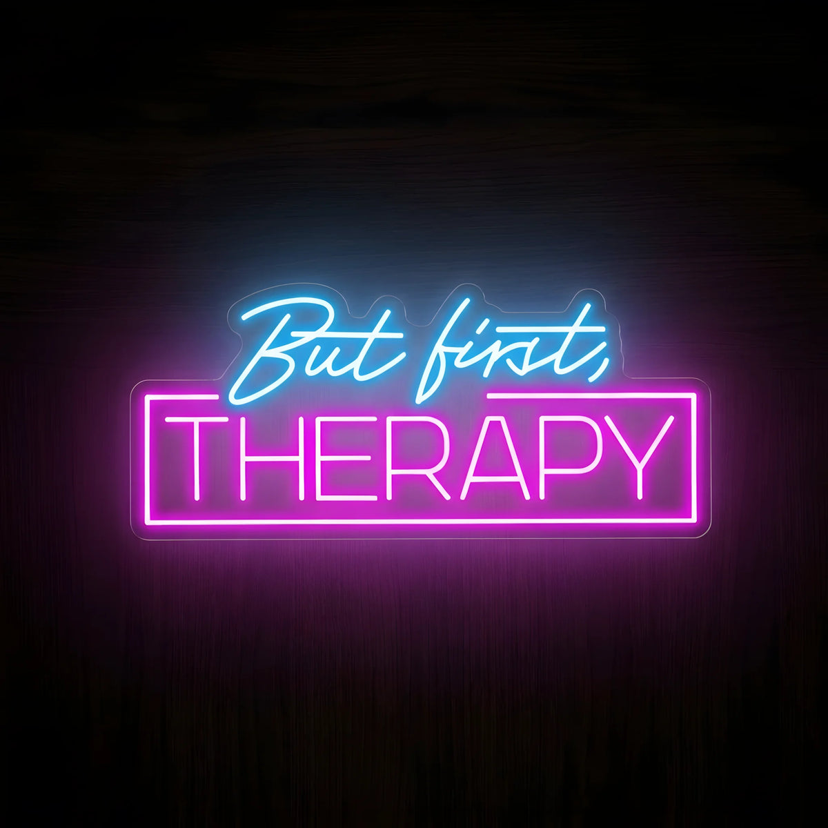 But First Therapy LED Neon Sign featuring blue cursive lettering above bright pink THERAPY text inside a rectangular neon border on clear acrylic against a dark wooden wall.