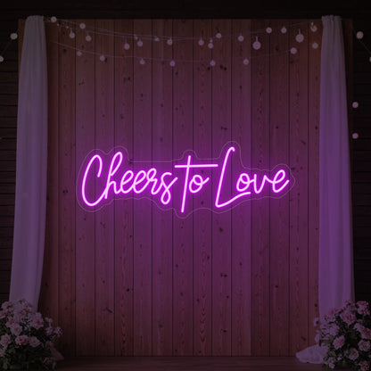 Photo of the 'Cheers To Love' neon sign glowing in deep pink against a styled interior wall.