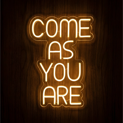 “COME AS YOU ARE” LED Neon Sign with warm white lettering on clear acrylic backing, glowing against a dark wood background and suited to living rooms, entryways, or reception areas.