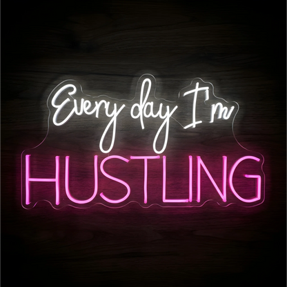 “Every day I’m HUSTLING” LED Neon Sign with white script lettering above bold pink uppercase text on clear acrylic against a dark wood background, suited to home offices and creative workspaces.