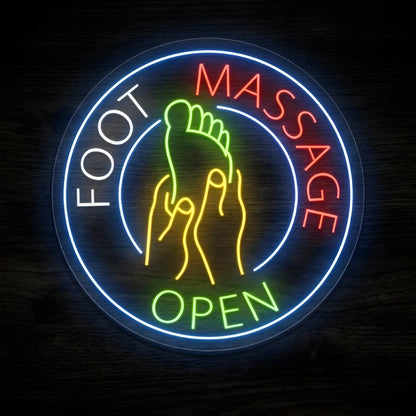 Foot Massage Open LED Neon Sign featuring a green foot being massaged by yellow hands, white FOOT lettering, red MASSAGE lettering, green OPEN text, and a glowing blue circular border against a dark wooden wall.