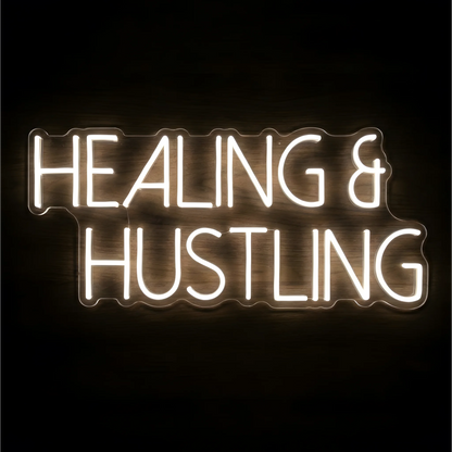“Healing & Hustling” LED Neon Sign glowing in bright white uppercase lettering on clear acrylic against a dark wood background, suited to home offices and creative workspaces.