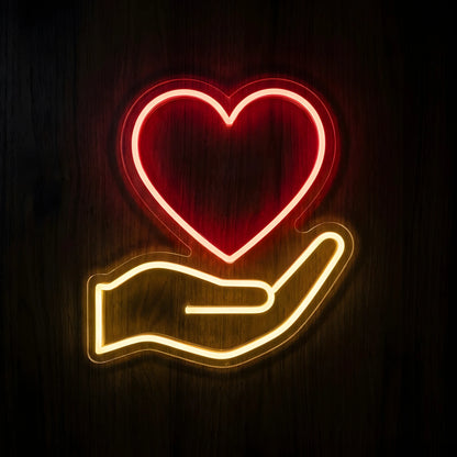 Healthcare Heart in Hand LED Neon Sign featuring a glowing red heart above a warm yellow open hand on clear cut-to-shape acrylic backing against a dark wooden wall.