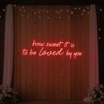 Photo of the 'How Sweet It Is To Be Loved By You' neon sign glowing in red against a styled interior wall.