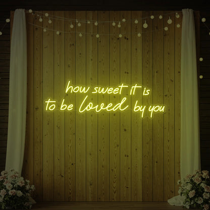 Photo of the 'How Sweet It Is To Be Loved By You' neon sign glowing in lemon yellow against a styled interior wall.