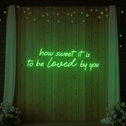 Photo of the 'How Sweet It Is To Be Loved By You' neon sign glowing in green against a styled interior wall.