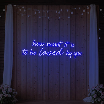 Photo of the 'How Sweet It Is To Be Loved By You' neon sign glowing in deep blue against a styled interior wall.