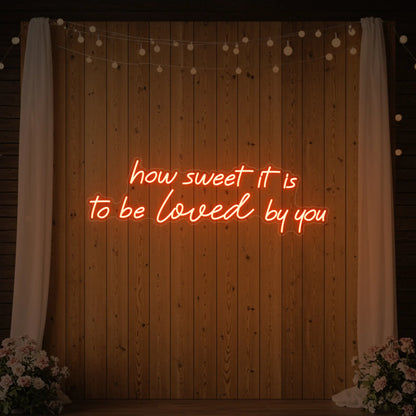 Photo of the 'How Sweet It Is To Be Loved By You' neon sign glowing in golden yellow against a styled interior wall.