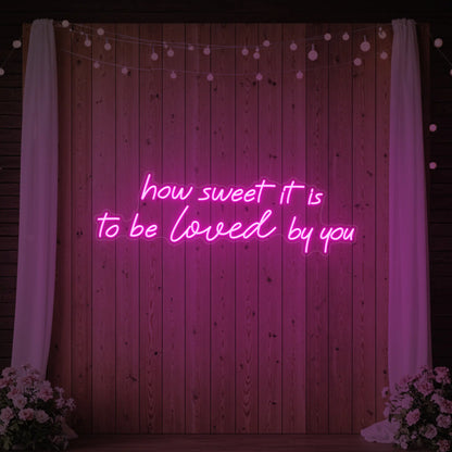 Photo of the 'How Sweet It Is To Be Loved By You' neon sign glowing in light pink against a styled interior wall.