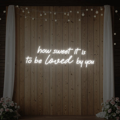 Photo of the 'How Sweet It Is To Be Loved By You' neon sign glowing in cool white against a styled interior wall.