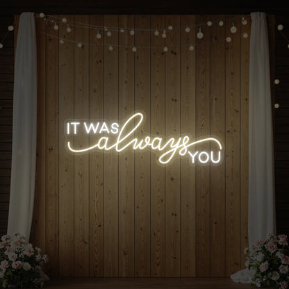 Photo of the 'It Was Always You' neon sign glowing in warm white against a styled interior wall.