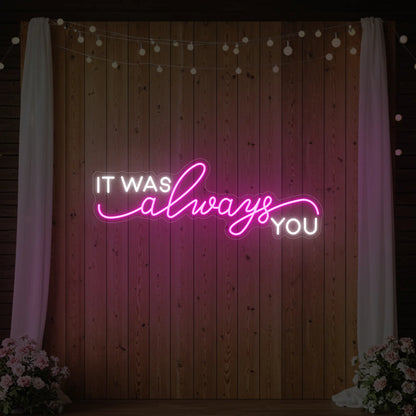 Photo of the 'It Was Always You' neon sign glowing in light pink against a styled interior wall.
