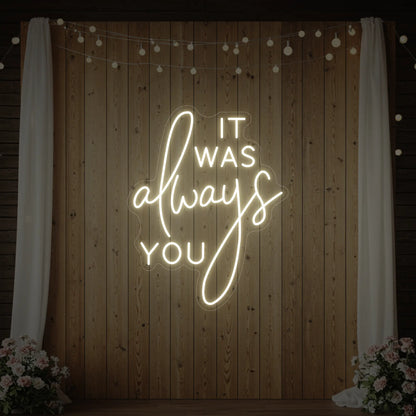 Photo of the 'It Was Always You' neon sign glowing in warm white against a styled interior wall.