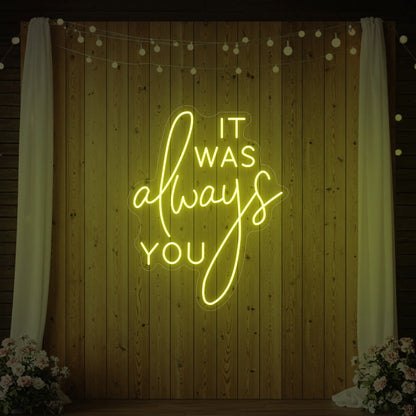 Photo of the 'It Was Always You' neon sign glowing in lemon yellow against a styled interior wall.