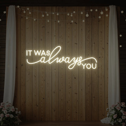 Photo of the 'It Was Always You' neon sign glowing in warm white against a styled interior wall.