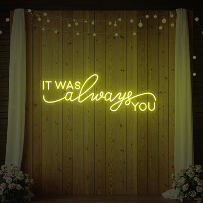 Photo of the 'It Was Always You' neon sign glowing in lemon yellow against a styled interior wall.