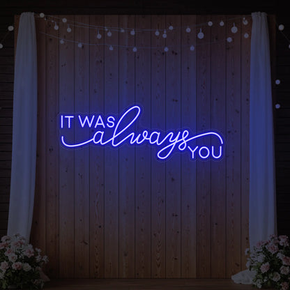 Photo of the 'It Was Always You' neon sign glowing in deep blue against a styled interior wall.