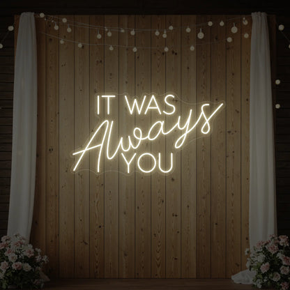 Photo of the 'It Was Always You' neon sign glowing in warm white against a styled interior wall.