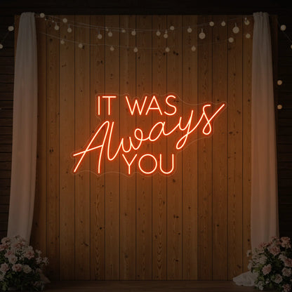Photo of the 'It Was Always You' neon sign glowing in golden yellow against a styled interior wall.