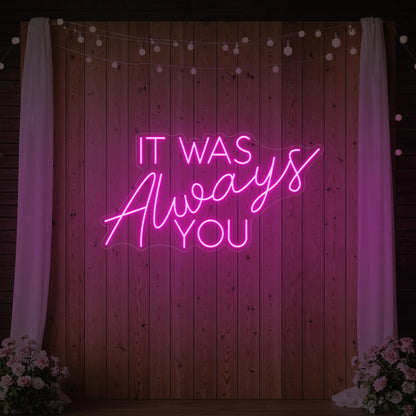 Photo of the 'It Was Always You' neon sign glowing in light pink against a styled interior wall.