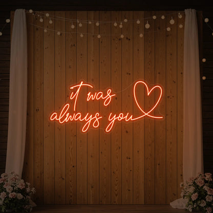 Photo of the 'It Was Always You Heart' neon sign glowing in golden yellow against a styled interior wall.