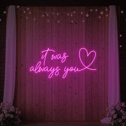 Photo of the 'It Was Always You Heart' neon sign glowing in light pink against a styled interior wall.