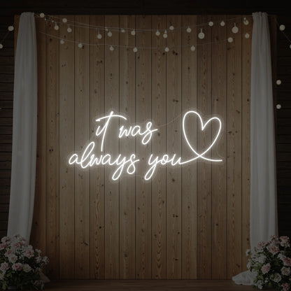 Photo of the 'It Was Always You Heart' neon sign glowing in cool white against a styled interior wall.
