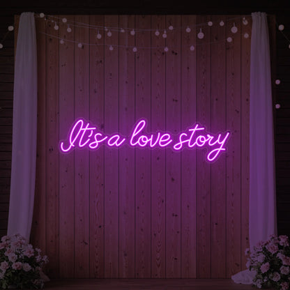 Photo of the 'It’s a Love Story' neon sign glowing in deep pink against a styled interior wall.
