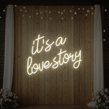 Photo of the 'It’s a Love Story' neon sign glowing in warm white against a styled interior wall.