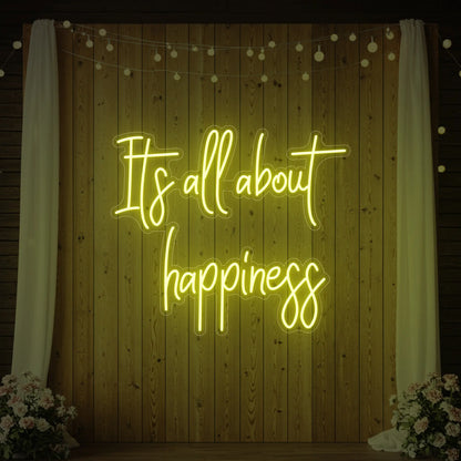 Photo of the 'It’s All About Happiness' neon sign glowing in lemon yellow against a styled interior wall.
