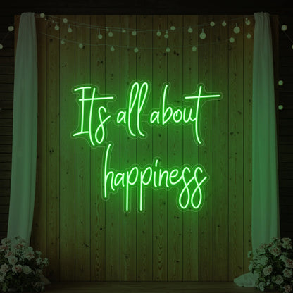 Photo of the 'It’s All About Happiness' neon sign glowing in green against a styled interior wall.