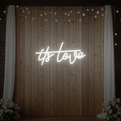 Photo of the 'It’s Love' neon sign glowing in cool white against a styled interior wall.