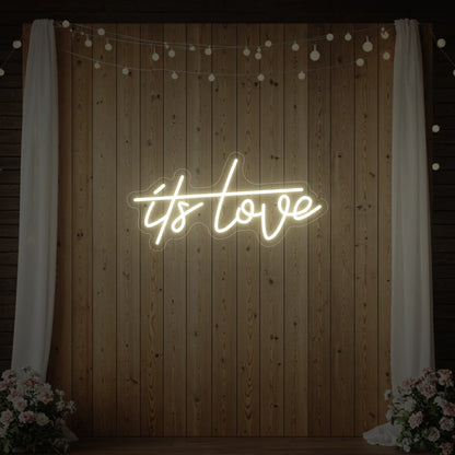 Photo of the 'It’s Love' neon sign glowing in warm white against a styled interior wall.