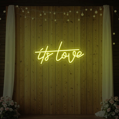 Photo of the 'It’s Love' neon sign glowing in lemon yellow against a styled interior wall.