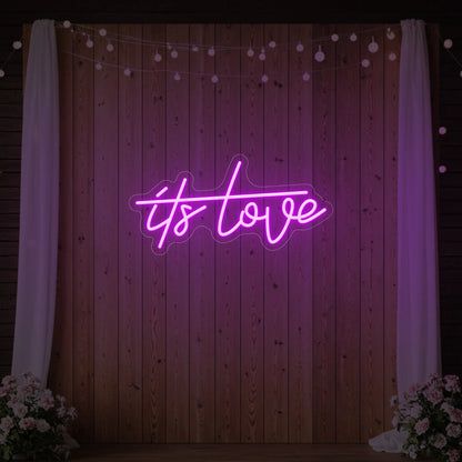 Photo of the 'It’s Love' neon sign glowing in deep pink against a styled interior wall.