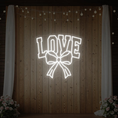 Photo of the 'Love and Ribbon' neon sign glowing in cool white against a styled interior wall.