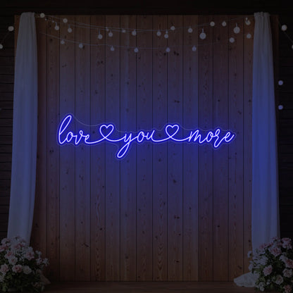 Photo of the 'Love You More' neon sign glowing in deep blue against a styled interior wall.