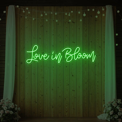 Photo of the 'Love in Bloom' neon sign glowing in green against a styled interior wall.