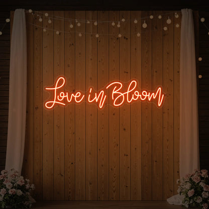 Photo of the 'Love in Bloom' neon sign glowing in golden yellow against a styled interior wall.