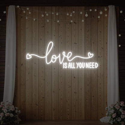 Photo of the 'Love Is All You Need' neon sign glowing in cool white against a styled interior wall.