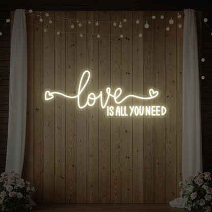 Photo of the 'Love Is All You Need' neon sign glowing in warm white against a styled interior wall.