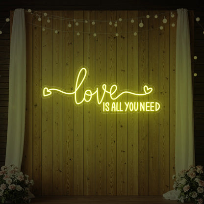 Photo of the 'Love Is All You Need' neon sign glowing in lemon yellow against a styled interior wall.